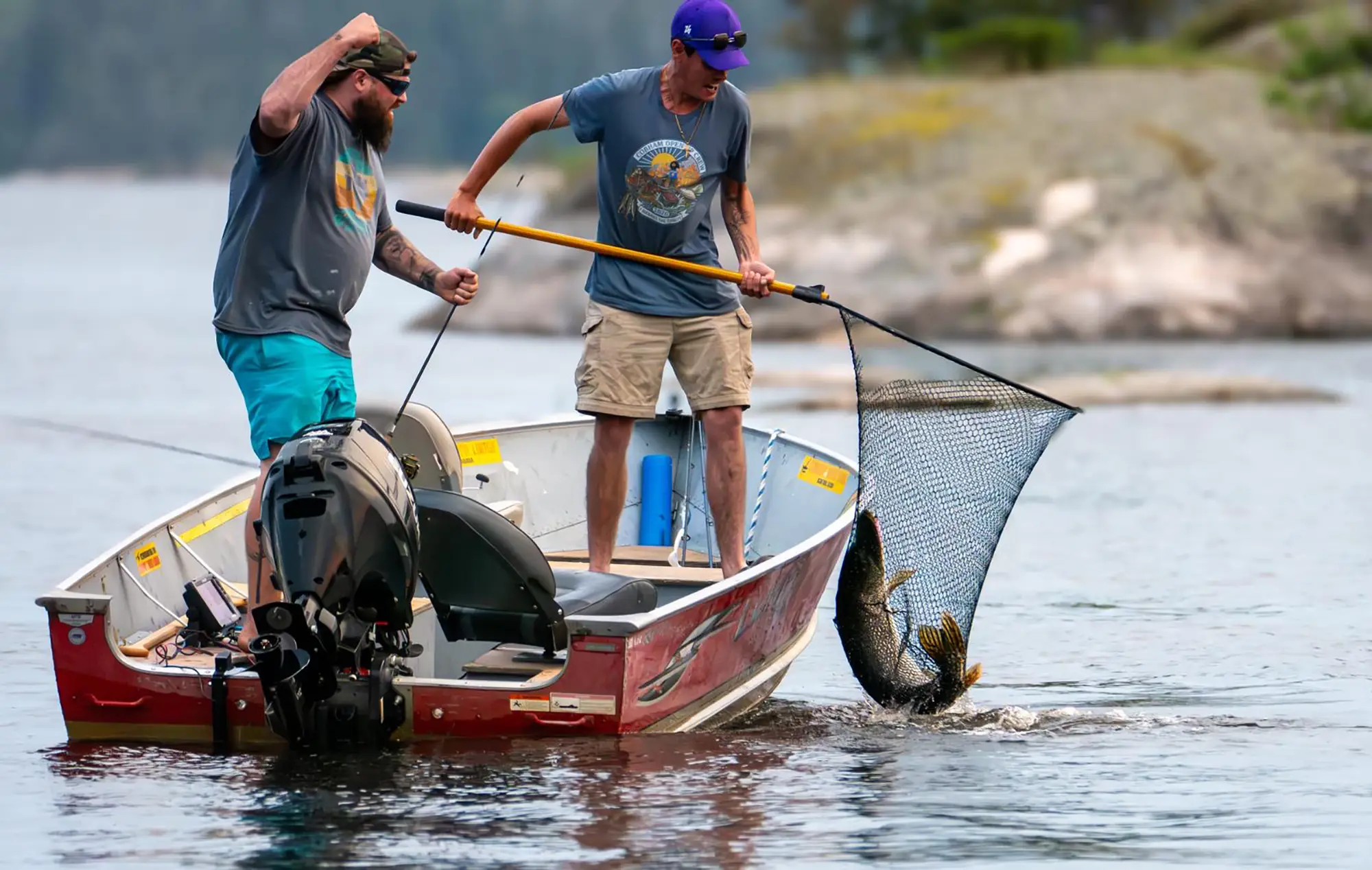 Two excited anglers in a red and aluminum fishing boat netting a large Northern Pike out of the water, with one cheering with a raised fist at Cobham River Lodge.