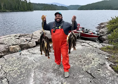 A smiling angler wearing bright orange bibs standing on a rocky shore, proudly holding two heavy stringers of freshly caught walleye with a moored fishing boat and forested lake in the background at Cobham River Lodge.