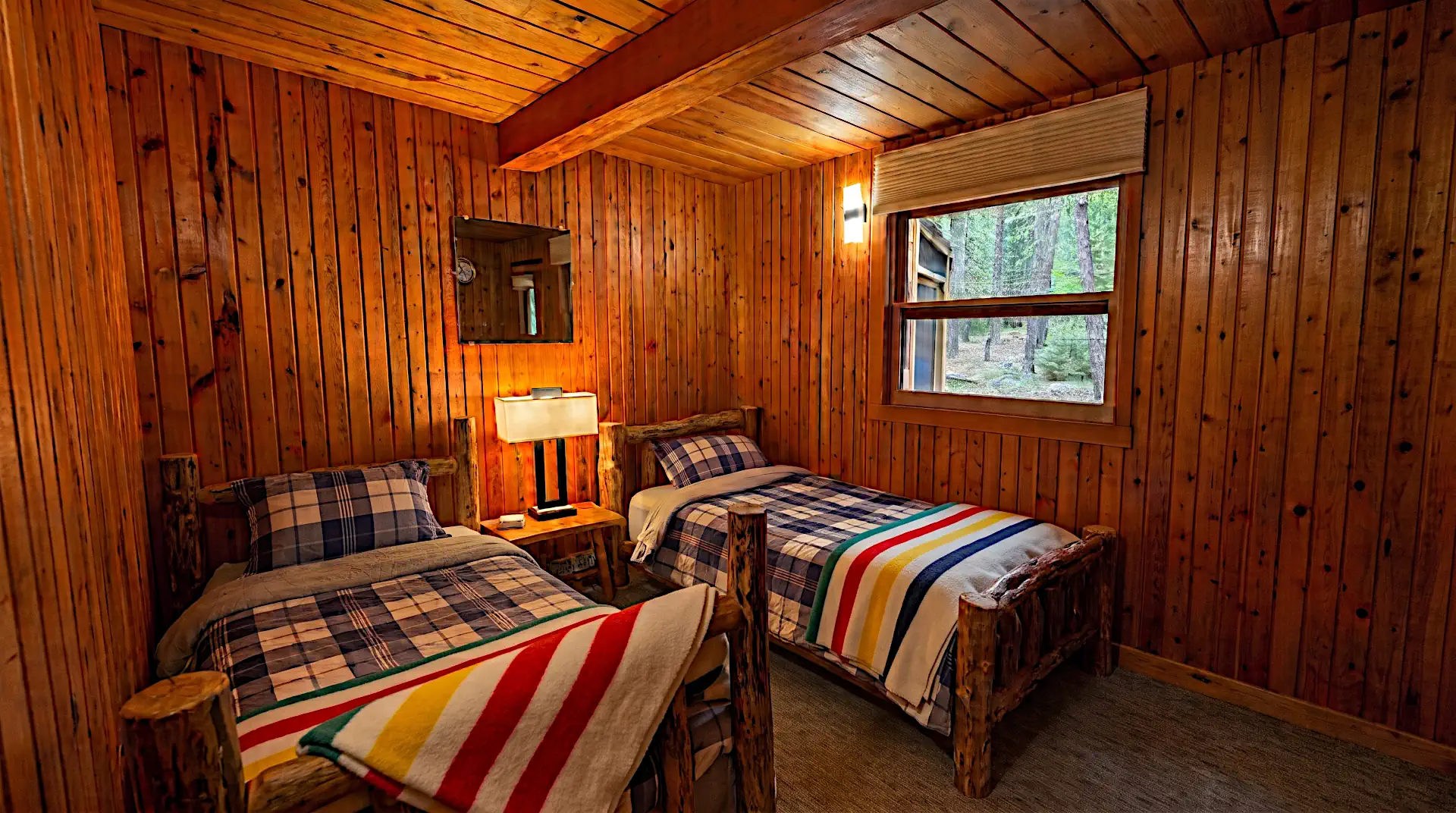 Alt Description: A cozy rustic cabin bedroom at Cobham River Lodge featuring fully wood-paneled walls, two twin log beds with plaid bedding and striped wool blankets, and a window overlooking a green pine forest.