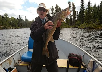 An excited female angler wearing a visor and dark jacket holding a large trophy Walleye vertically with its mouth open on a boat at Cobham River Outpost.