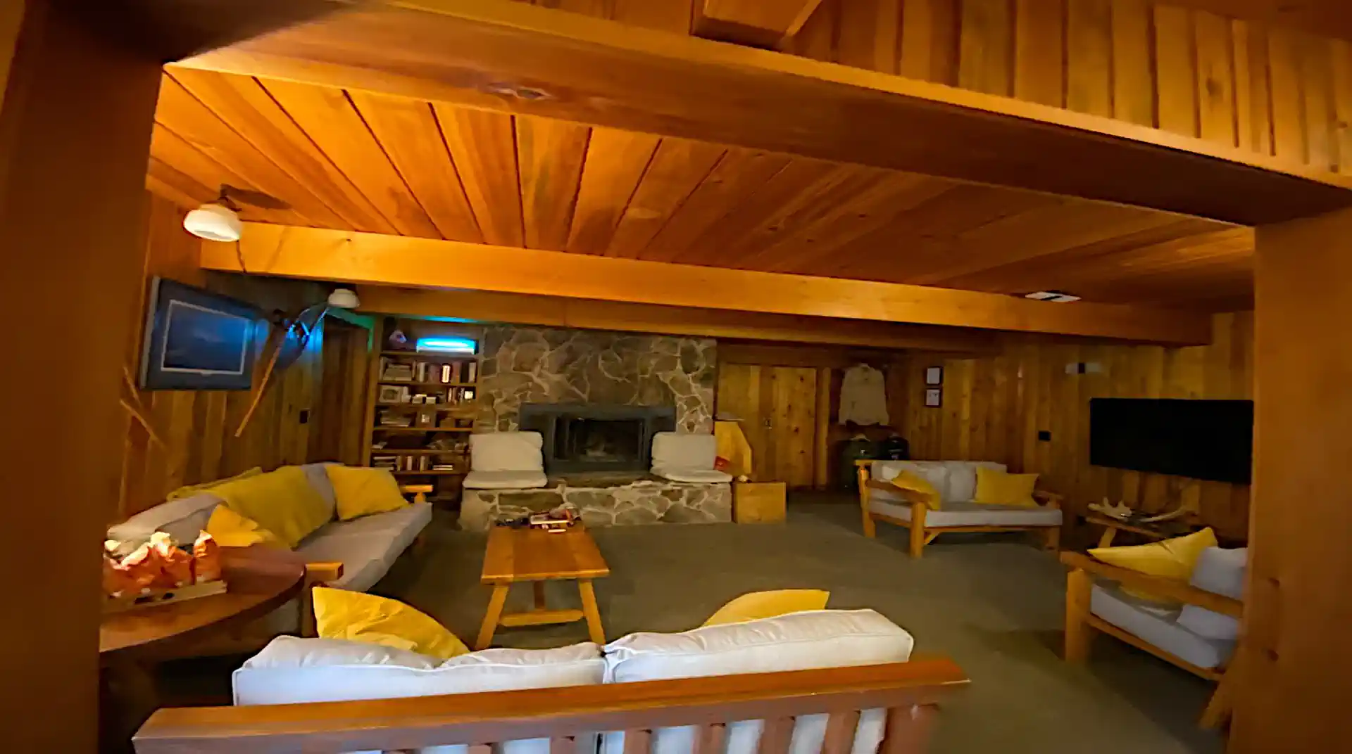 A spacious, T&G-paneled lounge area at Cobham River Lodge featuring exposed wooden beams, a large stone fireplace, and comfortable seating with bright yellow accent pillows.