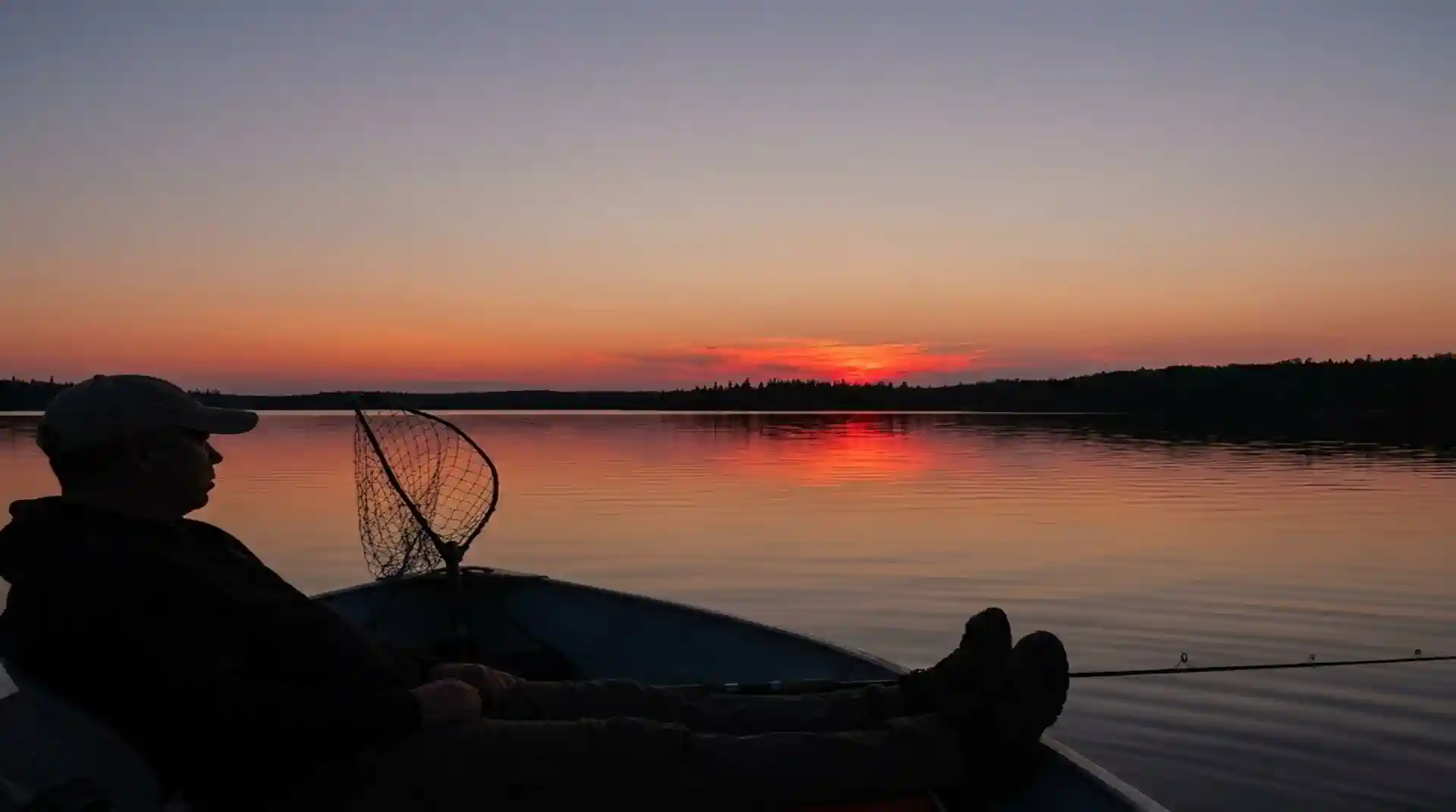 A silhouetted angler kicking back with feet up in a boat on calm, mirror-like lake water, watching a brilliant red and orange sunset over distant treelines at Cobham River Lodge.
