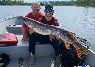 A smiling father and son sitting together in an aluminum boat, holding a massive, beautifully patterned trophy Northern Pike on a bright, sunny day at Cobham River Lodge.