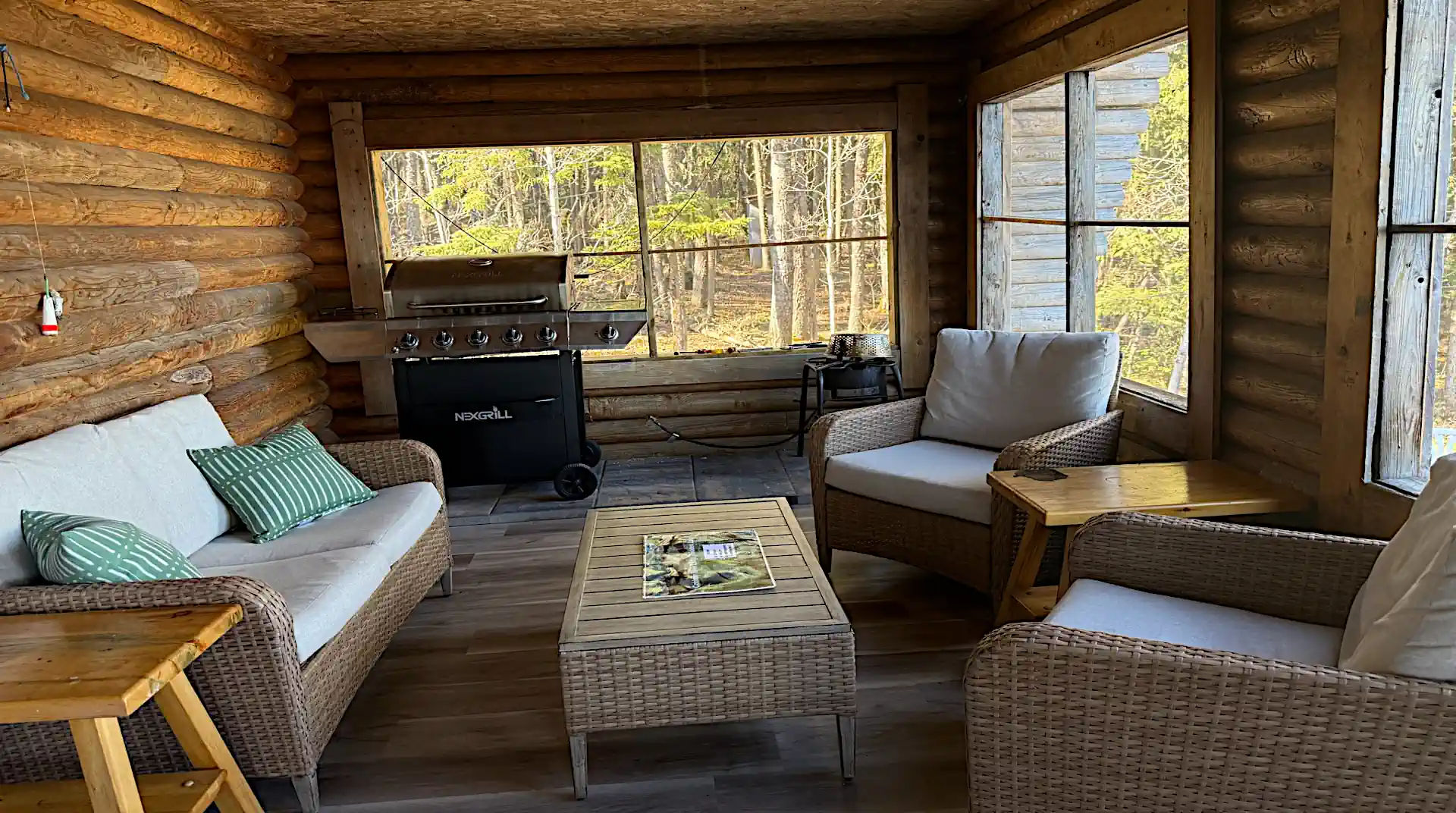 A cozy screened-in log porch featuring woven wicker seating with plush cushions, green striped throw pillows, coffee and end tables, and a large stainless steel Nexgrill BBQ grill at Cobham River Outpost.