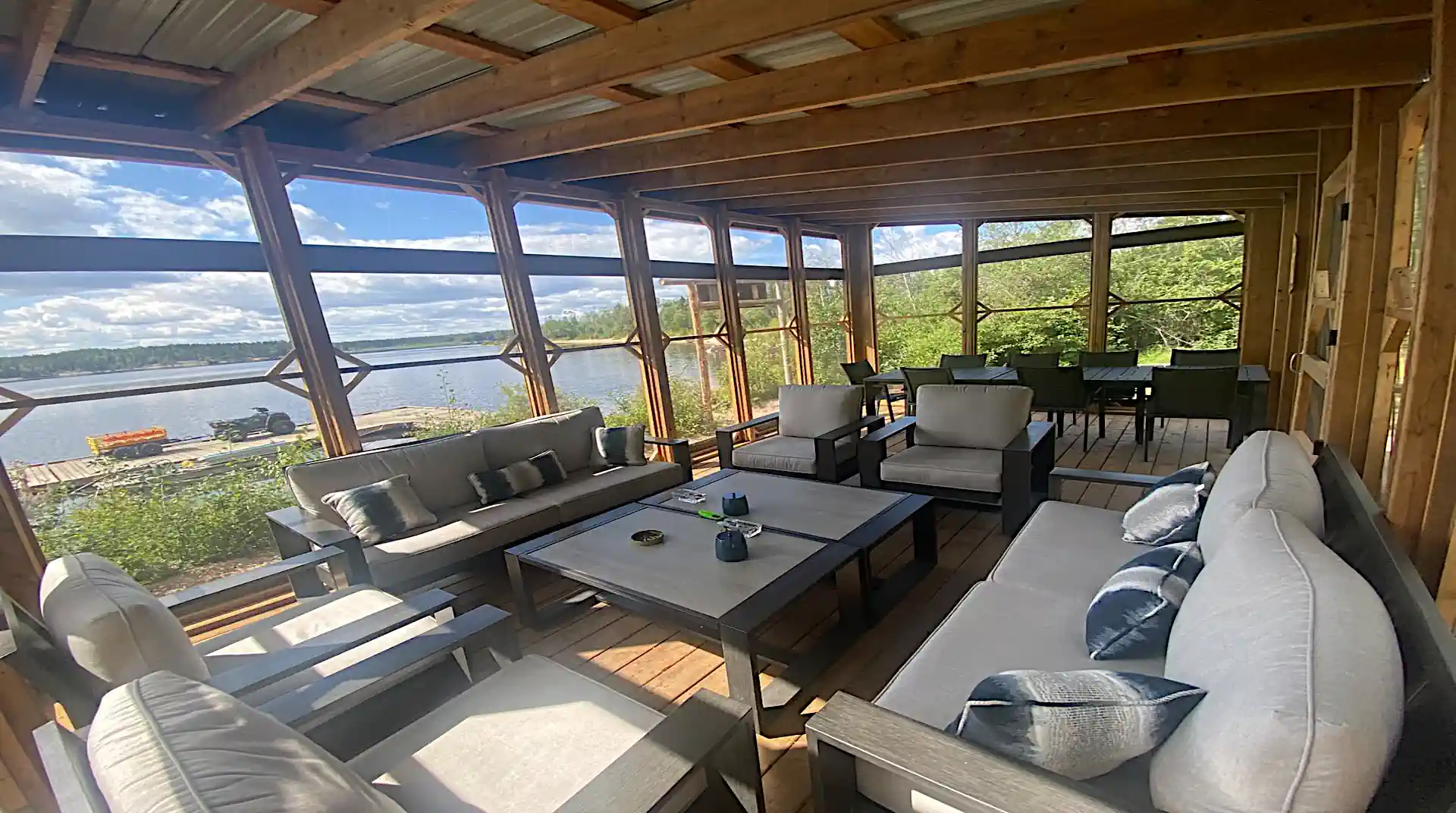 Alt Description: Interior of a spacious wooden screened-in gazebo equipped with comfortable cushioned outdoor sofas, armchairs, coffee tables, and dining seating overlooking the lake at Cobham River Lodge.