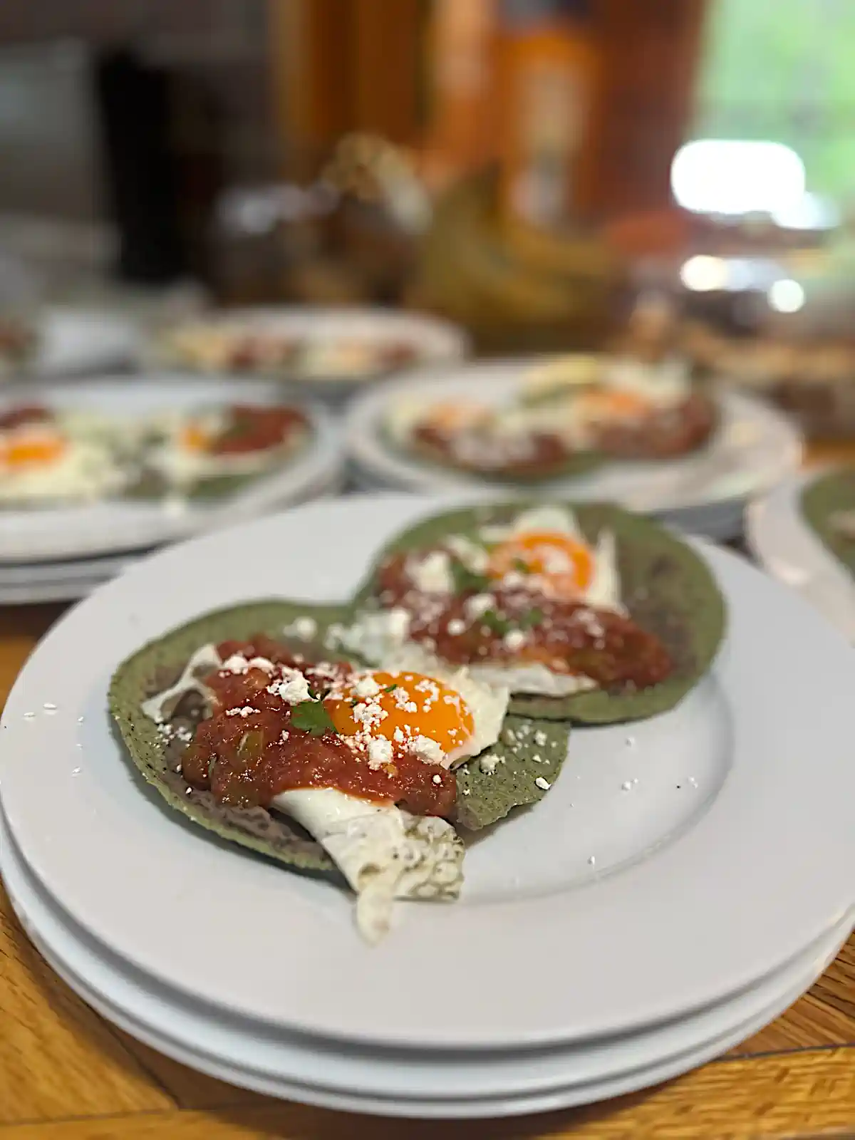 A close-up of a white plate with two huevos rancheros featuring fried eggs, salsa, and crumbly white cheese on green tortillas, with similar plates blurred in the background.