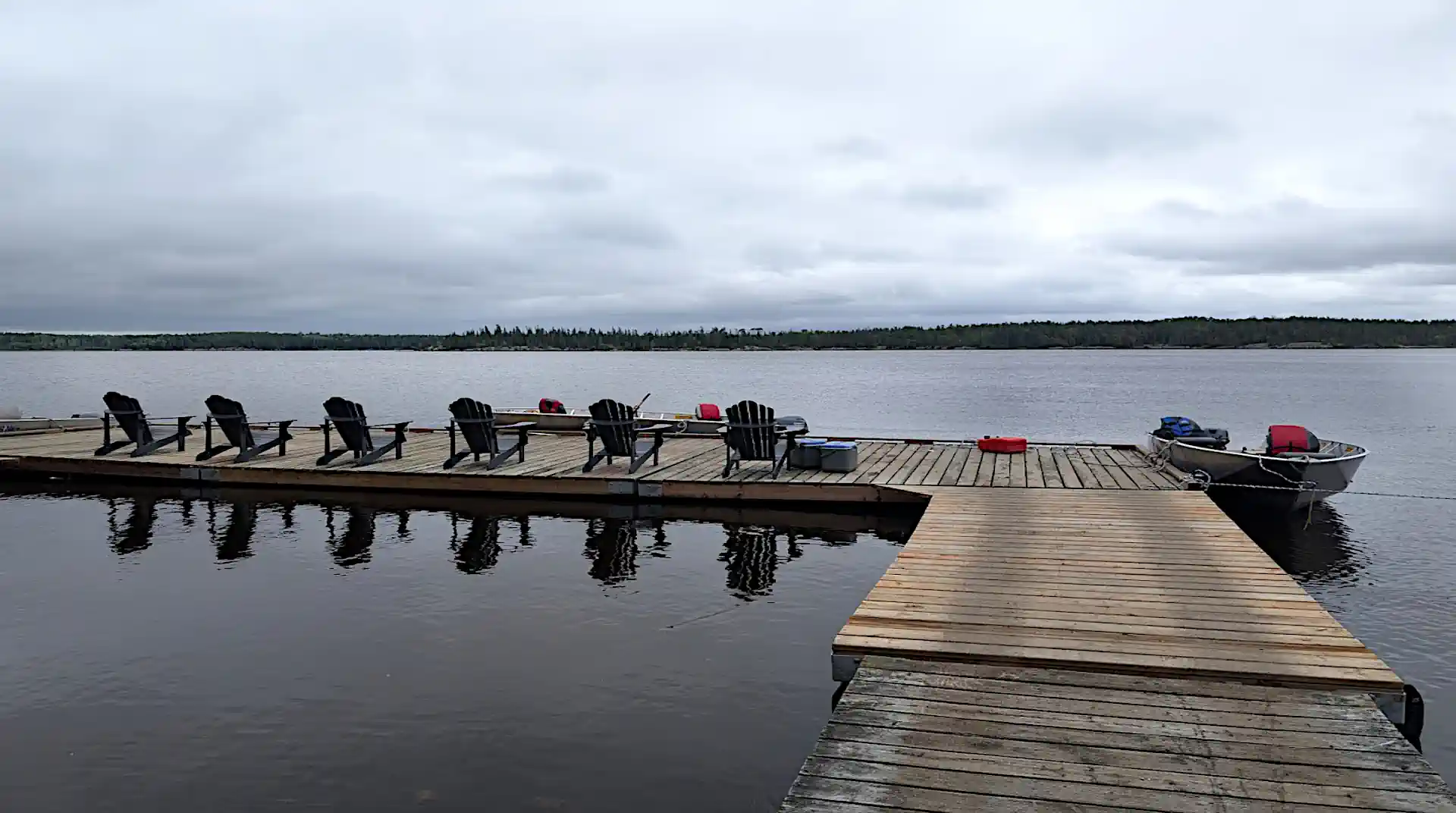 A long wooden dock extending into a calm river at Cobham River Lodge, featuring a row of six Adirondack chairs facing the water and an attached aluminum fishing boat under an overcast sky.