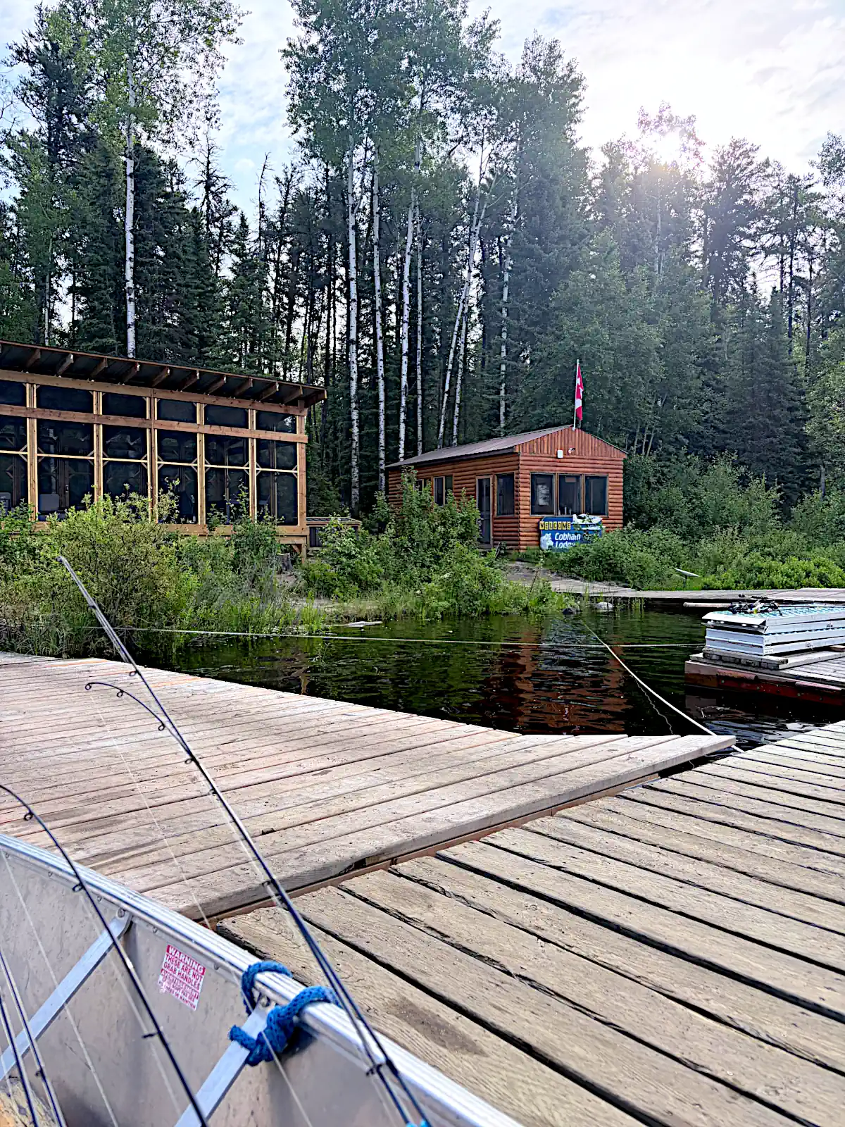 A view from a fishing boat at a wooden dock, looking toward the rustic fish house and lakeside screened-in viewing deck of Cobham River Lodge, nestled in a dense pine forest with a Canadian flag flying on the roof.