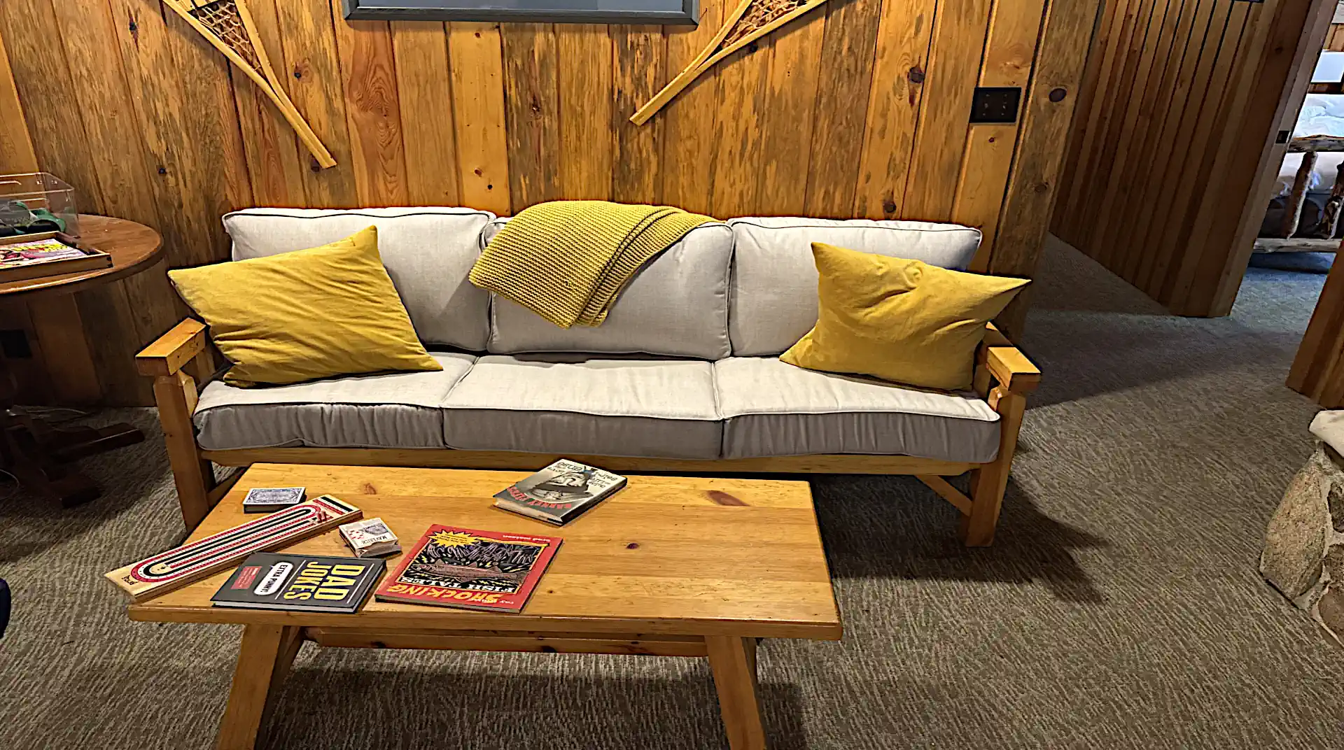 A cozy seating area at Cobham River Lodge featuring a light gray sofa with yellow pillows against a wood-paneled wall decorated with snowshoes, and a wooden coffee table holding books and a cribbage board.