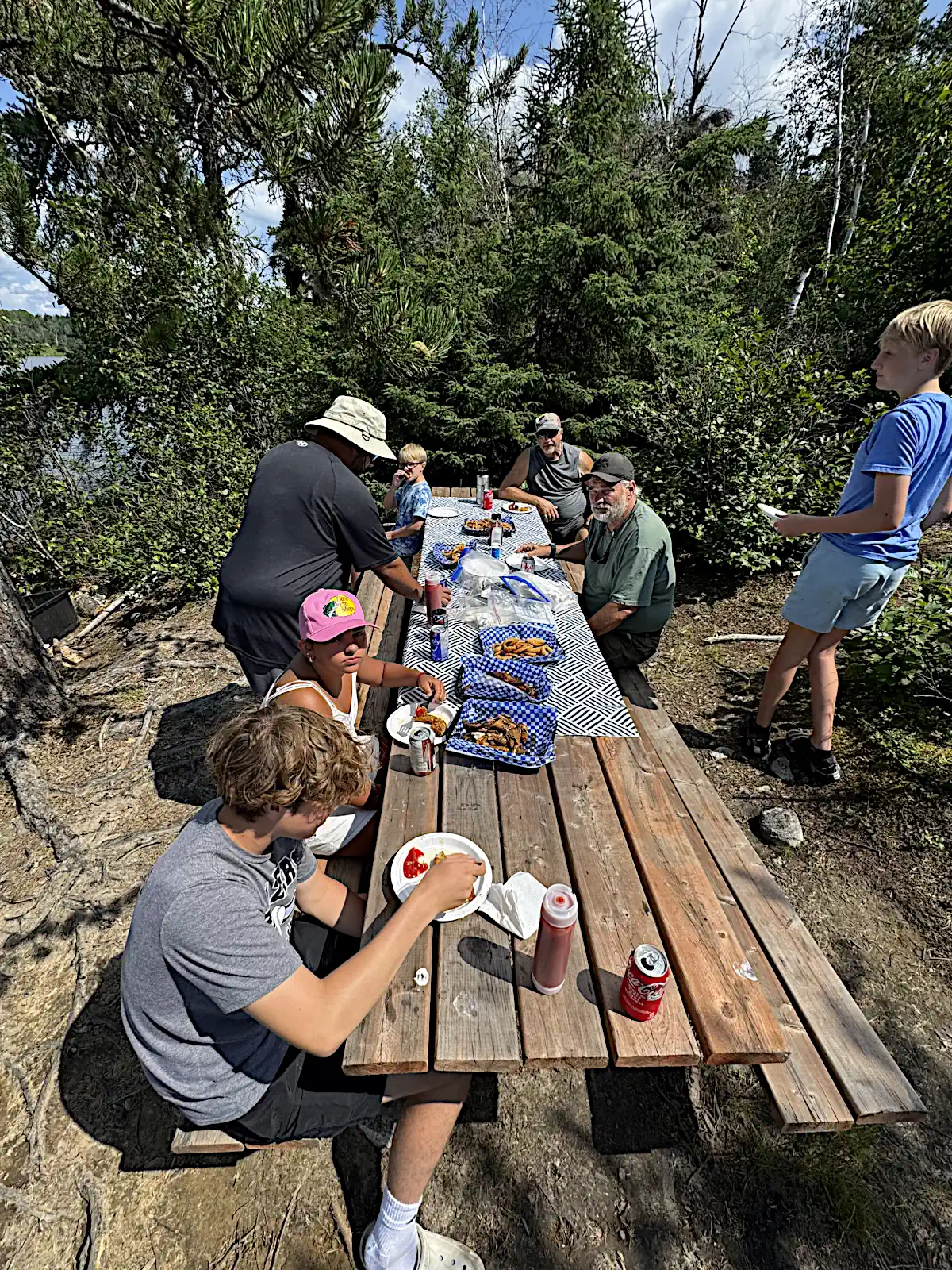 A group of guests sitting at a long wooden picnic table in a sunny wooded area, enjoying a shore lunch of fried food and drinks at Cobham River Lodge.