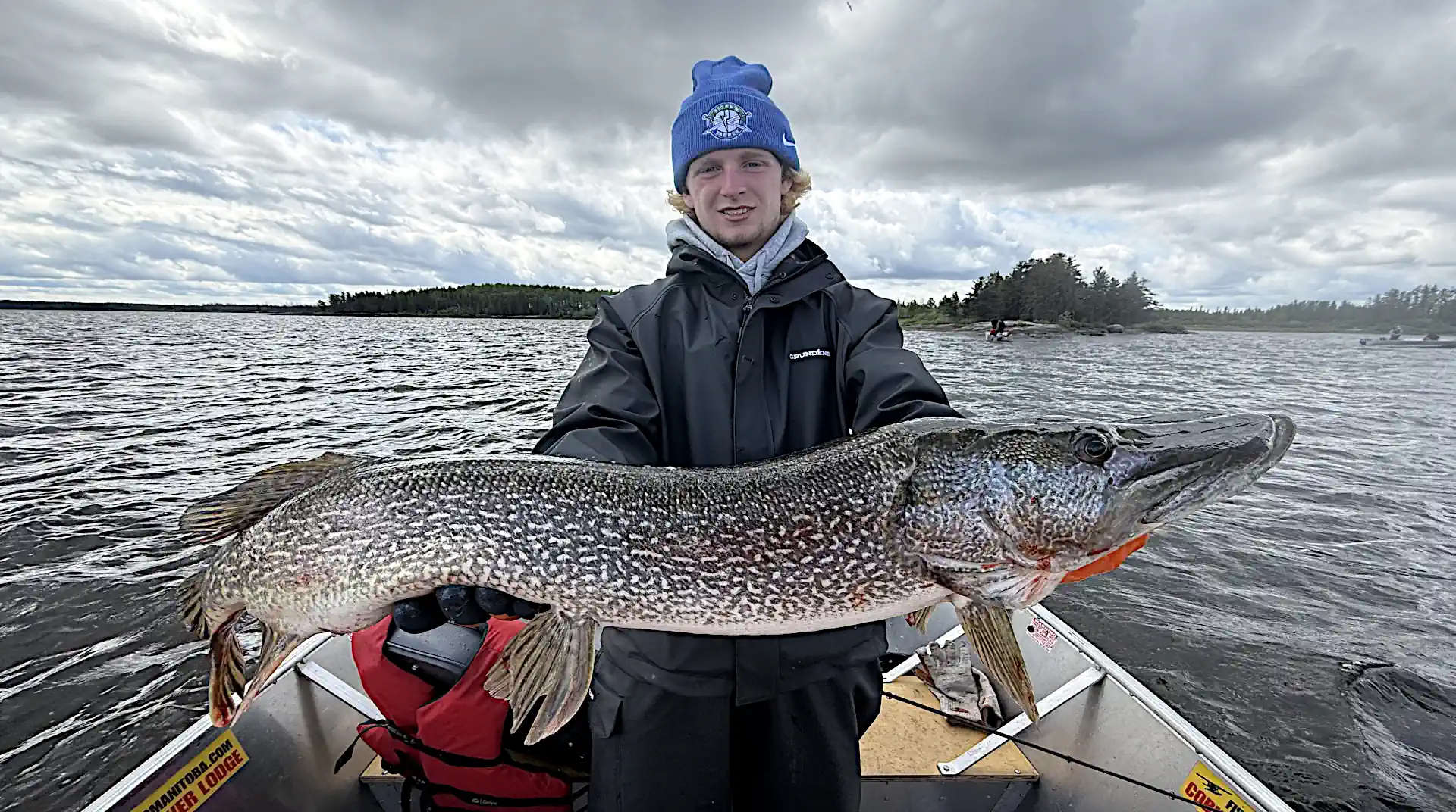 A young angler in a black waterproof jacket and blue beanie holding a massive trophy Northern Pike horizontally in an aluminum boat on open water at Cobham River Lodge.