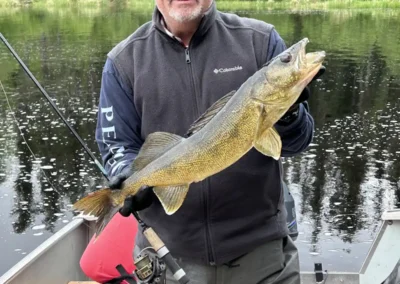 A smiling man in a fishing boat wearing a cap and dark fleece vest, proudly holding a large golden walleye horizontally with both gloved hands against a backdrop of a calm river and green pine forest at Cobham River Outpost.