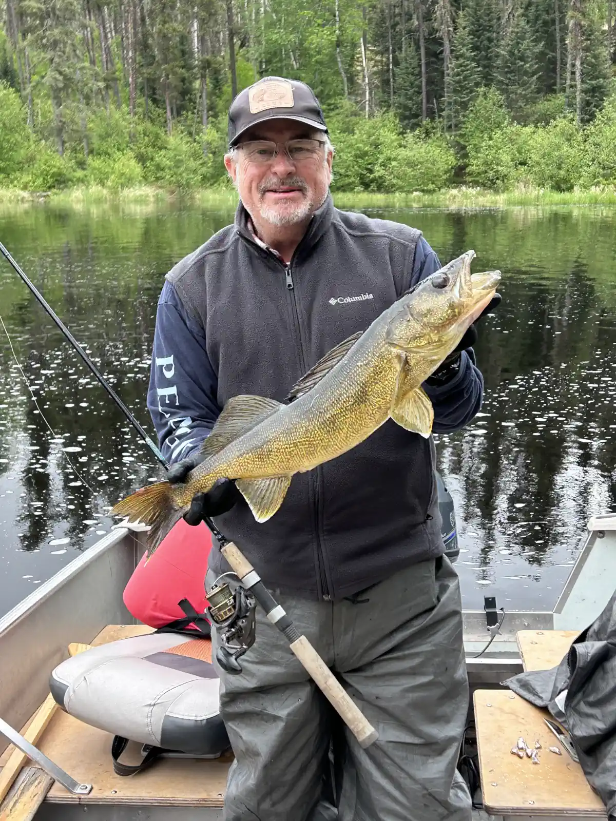 A smiling man in a fishing boat wearing a cap and dark fleece vest, proudly holding a large golden walleye horizontally with both gloved hands against a backdrop of a calm river and green pine forest at Cobham River Outpost.