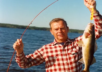 A man in a red and white checkered flannel shirt standing in a small boat on a sunny day, proudly holding up a freshly caught walleye on a yellow lure with a bent red fishing rod in his other hand at Cobham River Outpost.