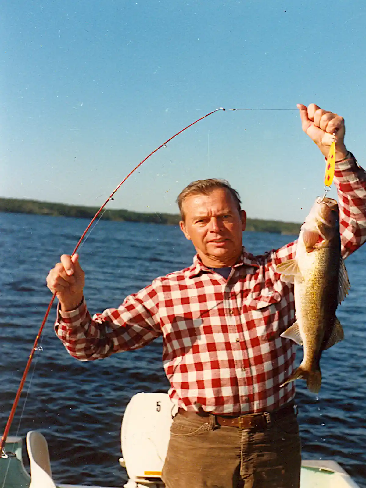A man in a red and white checkered flannel shirt standing in a small boat on a sunny day, proudly holding up a freshly caught walleye on a yellow lure with a bent red fishing rod in his other hand at Cobham River Outpost.