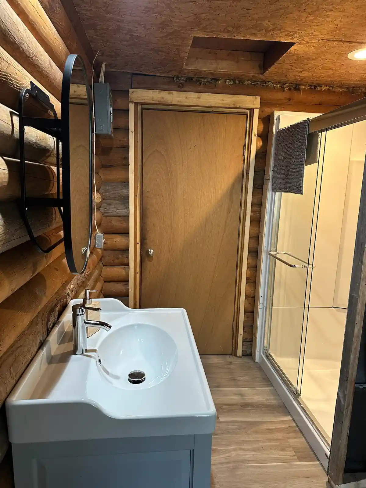 A rustic log cabin bathroom at Cobham River Outpost featuring exposed log walls, a modern white sink vanity, a round mirror, and a glass-enclosed shower.