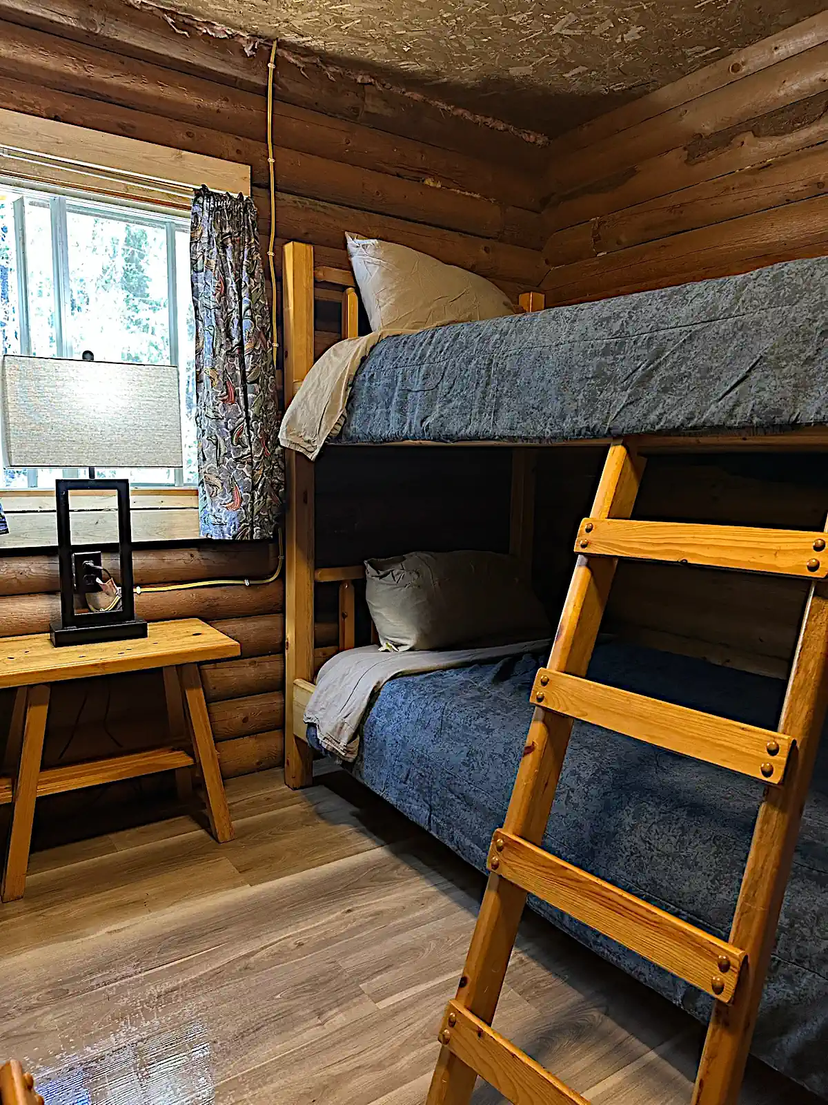 A cozy, rustic log cabin bedroom at Cobham River Outpost featuring a sturdy wooden bunk bed with a ladder, blue blankets, and a small wooden nightstand with a lamp next to a window.