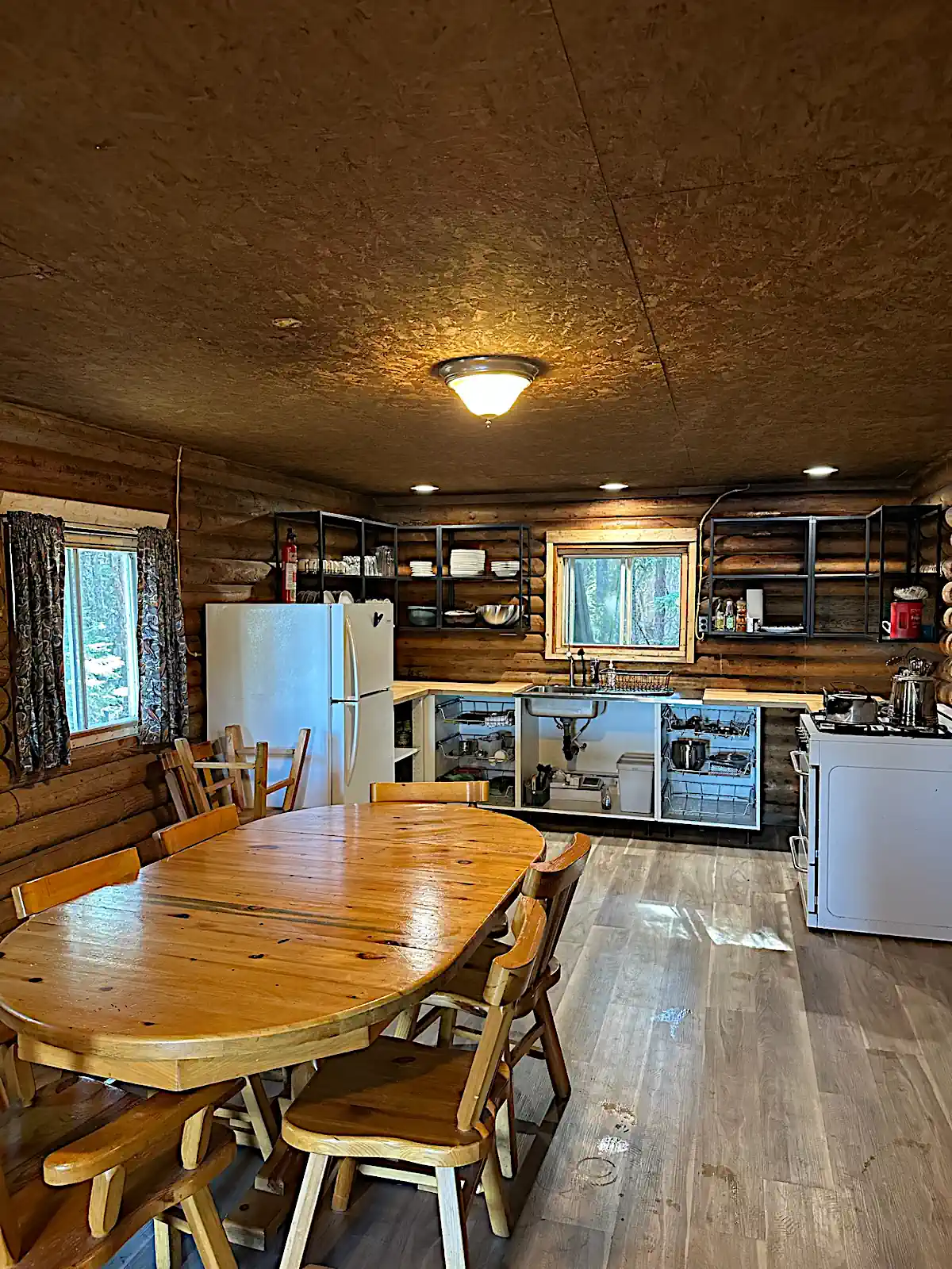 A spacious rustic log cabin interior at Cobham River Outpost featuring a large oval wooden dining table with chairs, seamlessly connected to a fully equipped kitchen with white appliances, open shelving, and exposed log walls.