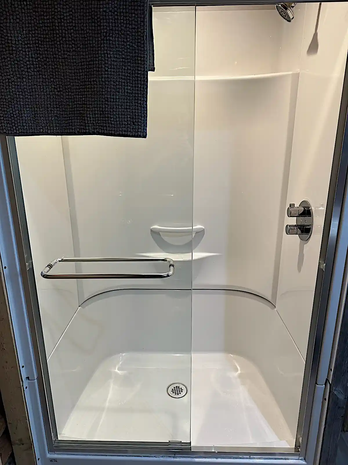A close-up view of a clean, modern white shower stall featuring a glass sliding door, chrome fixtures, and a grey towel draped over the top edge in a cabin bathroom at Cobham River Outpost.
