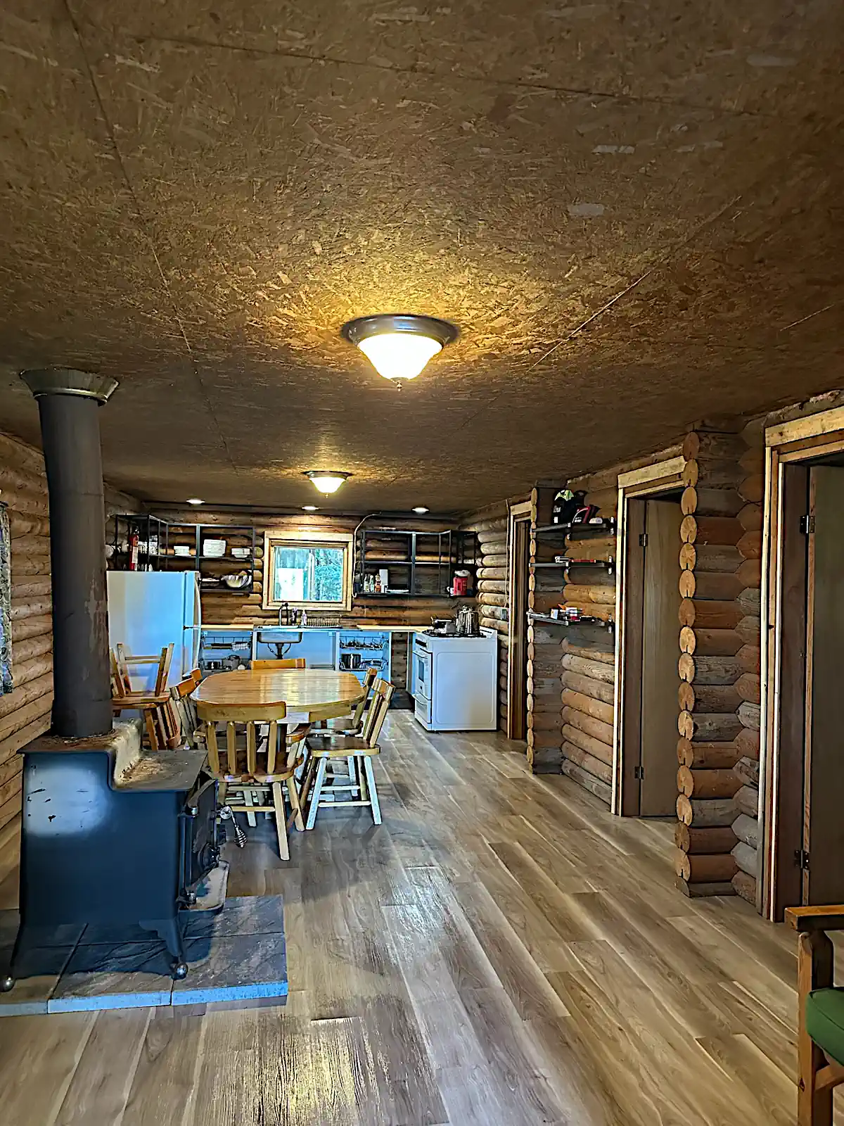 A wide interior view of a rustic log cabin at Cobham River Outpost, featuring exposed log walls, a black wood-burning stove, a wooden dining table, and a kitchen area with white appliances.