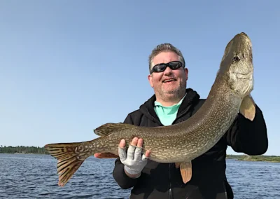 A smiling man wearing sunglasses and a black jacket standing on a boat, proudly holding a massive Northern Pike with gloved hands under a clear blue sky at Cobham River Outpost.