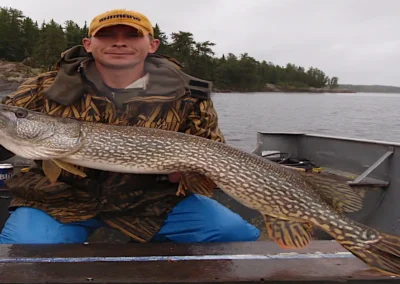 A smiling man in a yellow cap and camouflage jacket sitting in an aluminum boat, proudly holding a massive Northern Pike horizontally over a measuring board at Cobham River Outpost.