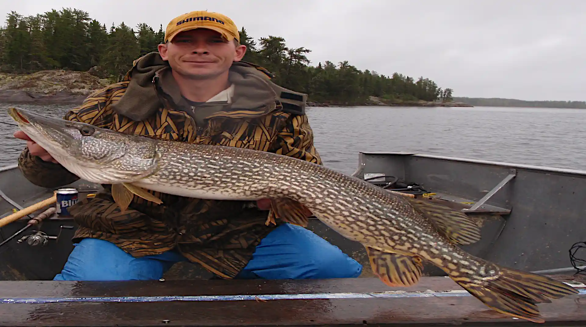 A smiling man in a yellow cap and camouflage jacket sitting in an aluminum boat, proudly holding a massive Northern Pike horizontally over a measuring board at Cobham River Outpost.