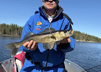A man wearing sunglasses and a bright blue Bass Pro Shops rain suit standing in an aluminum fishing boat, proudly holding a walleye with both hands on a sunny day at Cobham River Outpost.