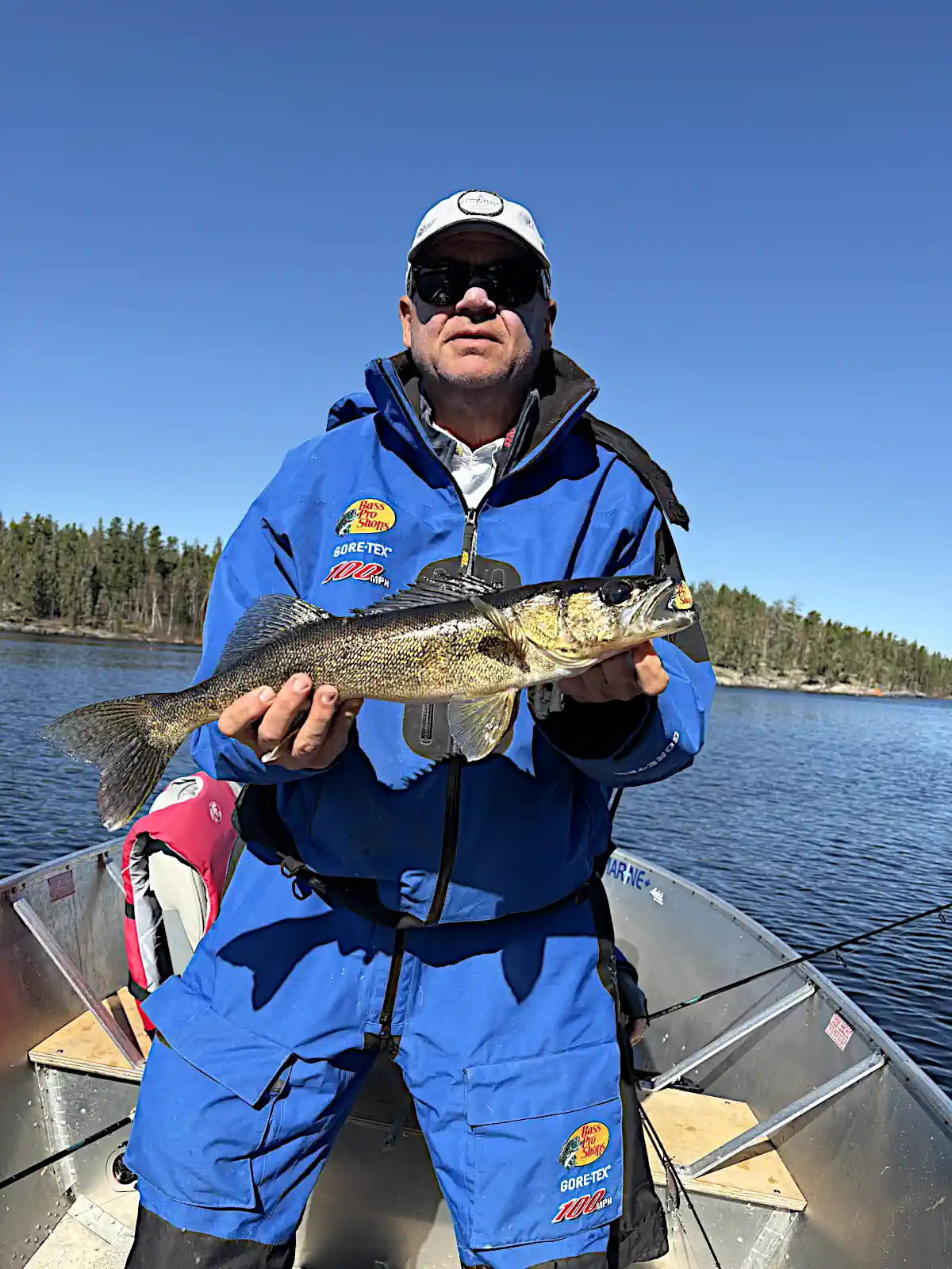 A man wearing sunglasses and a bright blue Bass Pro Shops rain suit standing in an aluminum fishing boat, proudly holding a walleye with both hands on a sunny day at Cobham River Outpost.