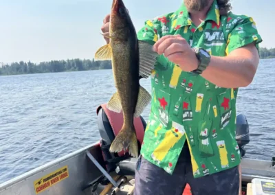 A bearded man wearing sunglasses and a bright green patterned shirt standing in an aluminum boat, holding a walleye vertically on a sunny day at Cobham River Outpost.