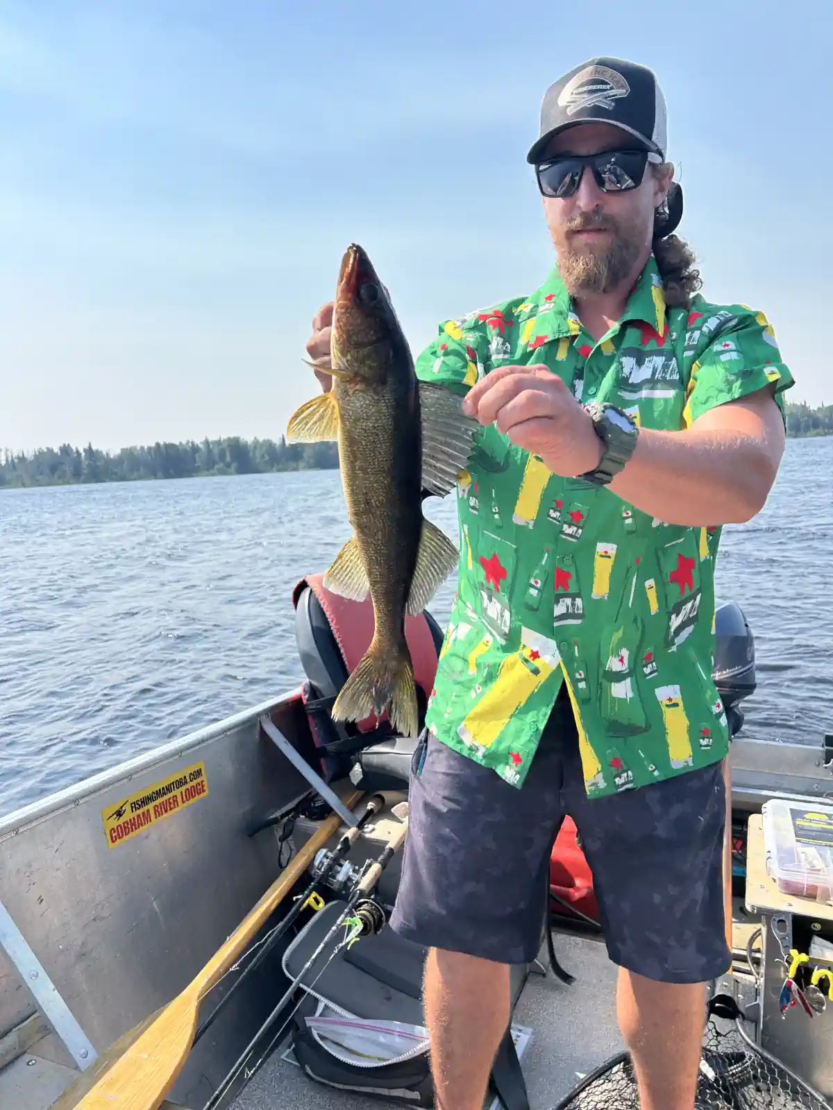 A bearded man wearing sunglasses and a bright green patterned shirt standing in an aluminum boat, holding a walleye vertically on a sunny day at Cobham River Outpost.