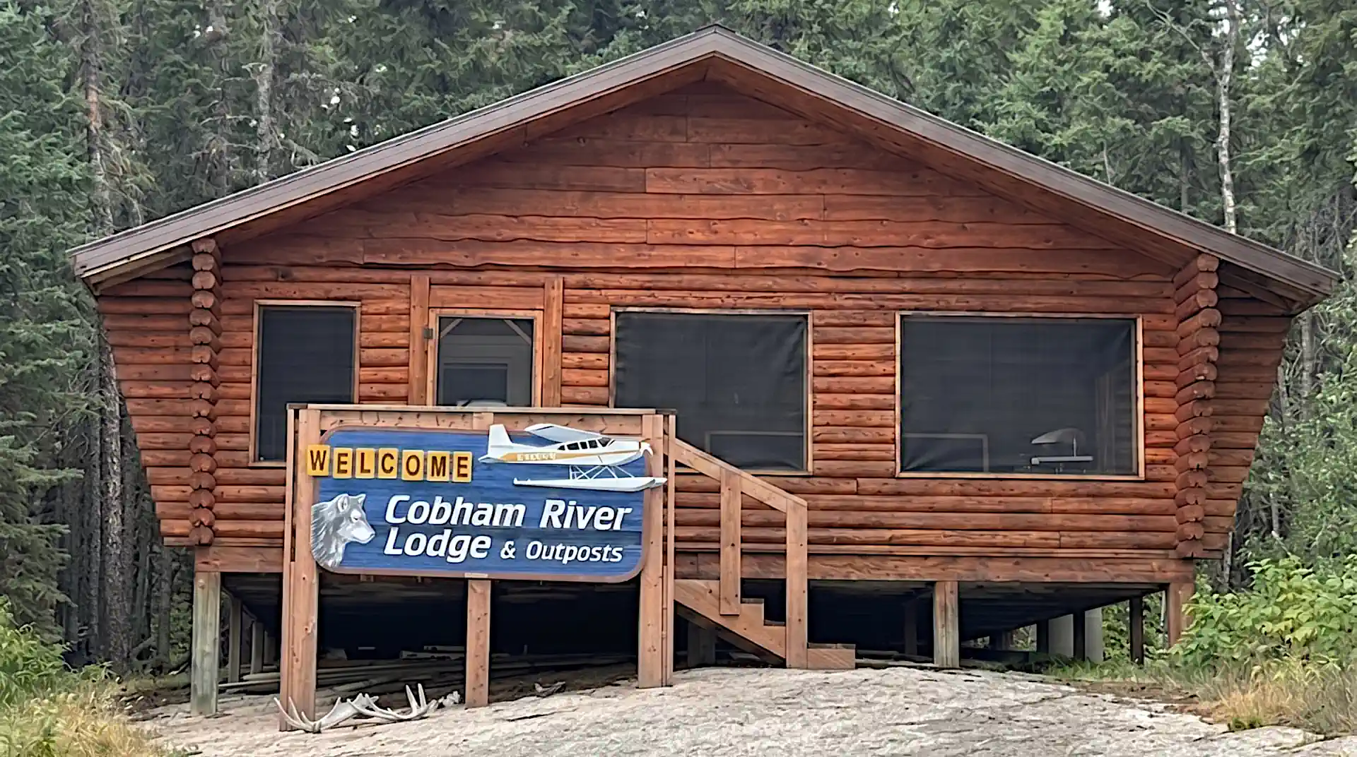 A classic wooden log cabin elevated on posts with a prominent blue "Welcome to Cobham River Lodge & Outposts" sign in the foreground, surrounded by a dense green pine forest