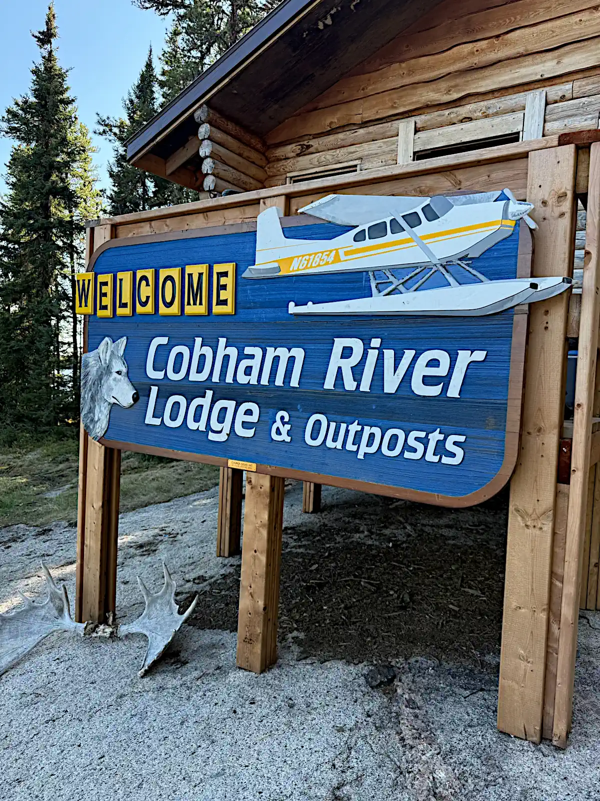 A close-up of a large blue wooden sign reading "Welcome Cobham River Lodge & Outposts," featuring cutout illustrations of a yellow and white floatplane and a wolf's head, standing in front of a log cabin with moose antlers on the rocky ground below.