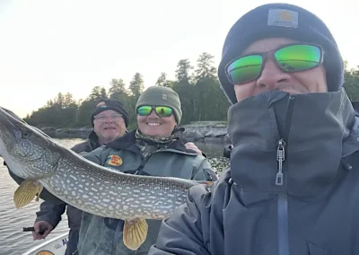 Fisherman holding a trophy Northern Pike with dense evergreen pine trees in the background in Manitoba.