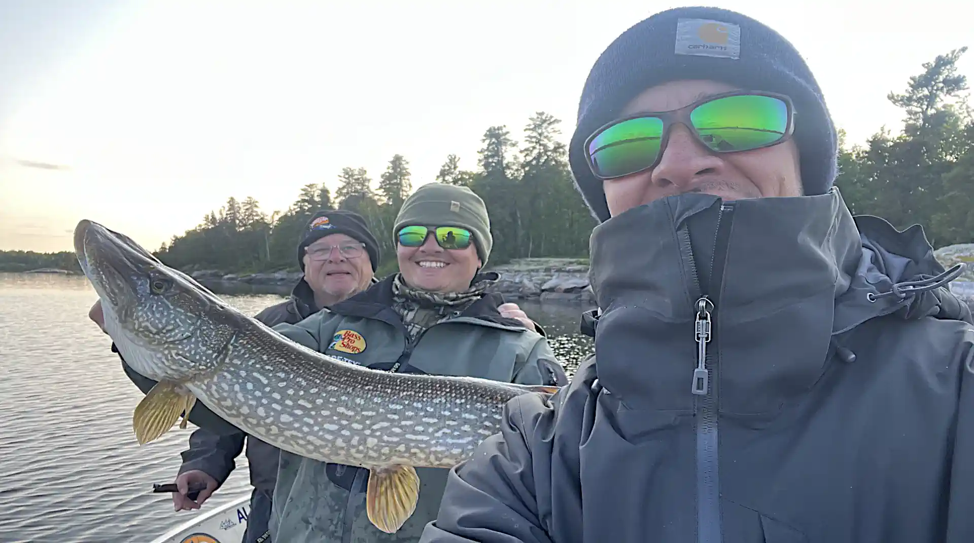 Fisherman holding a trophy Northern Pike with dense evergreen pine trees in the background in Manitoba.
