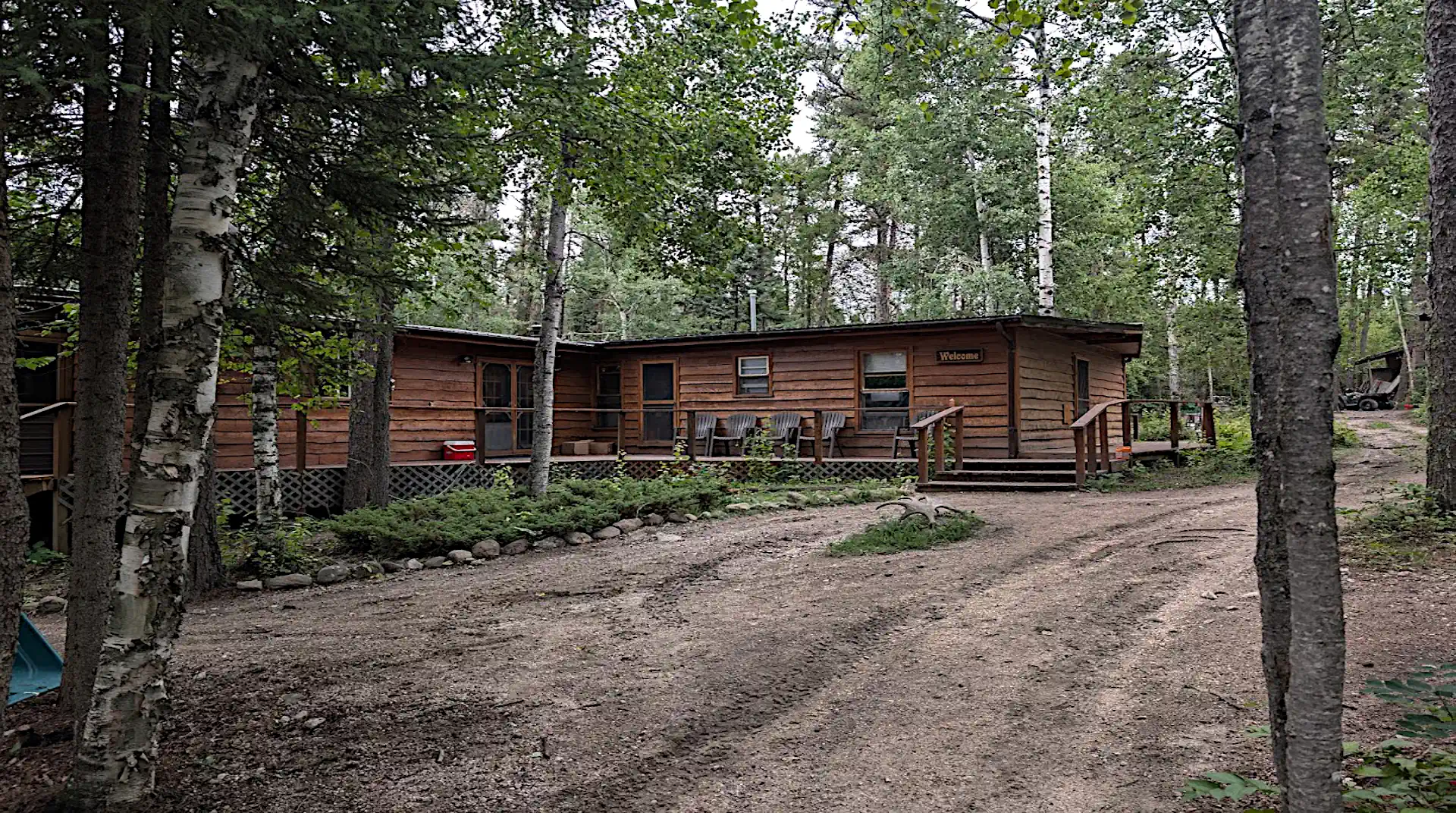 A rustic, T-shaped lodge with a large wooden deck and a "Welcome" sign, surrounded by birch and pine trees at Cobham River Lodge.